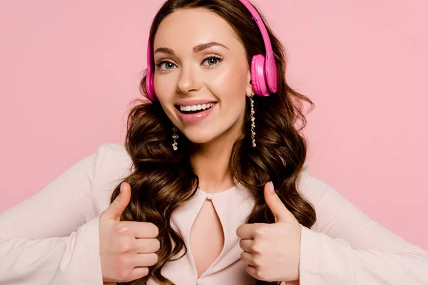 Alegre chica escuchando música en auriculares inalámbricos y mostrando los pulgares hacia arriba aislado en rosa - foto de stock