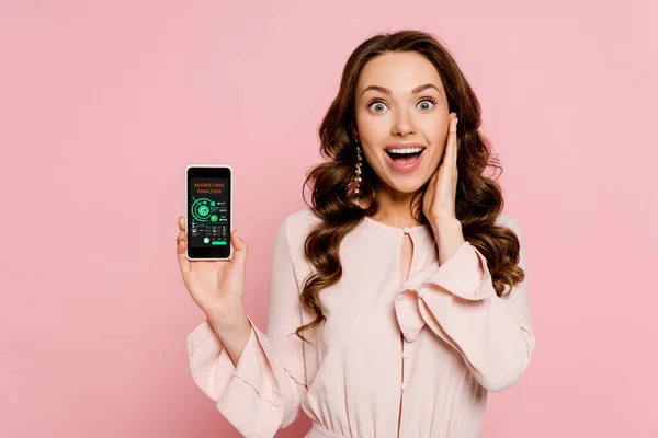 Aufgeregtes Mädchen mit Smartphone mit Diagrammen und Diagrammen auf dem Bildschirm und Blick auf Kamera isoliert auf rosa — Stockfoto