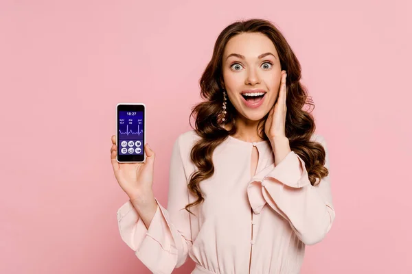 Chica emocionada sosteniendo el teléfono inteligente con aplicación médica en la pantalla y mirando a la cámara aislada en rosa - foto de stock