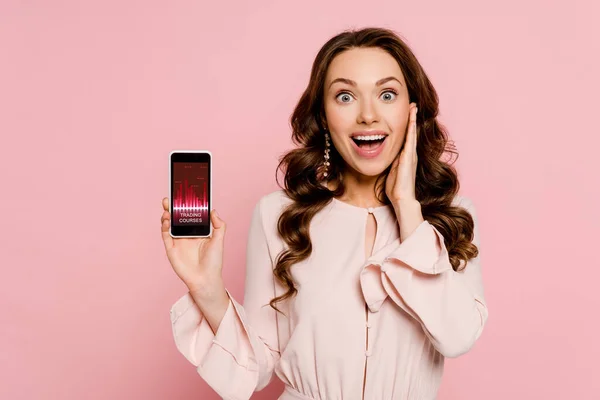 Excited girl holding smartphone with trading courses on screen and looking at camera isolated on pink — Stock Photo
