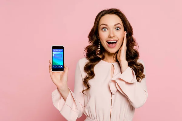 Excited girl holding smartphone with booking on screen and looking at camera isolated on pink — Stock Photo