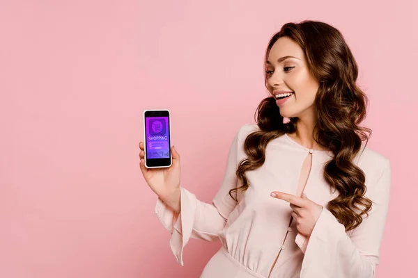 Alegre chica apuntando con el dedo en el teléfono inteligente con compras en línea en la pantalla aislado en rosa - foto de stock