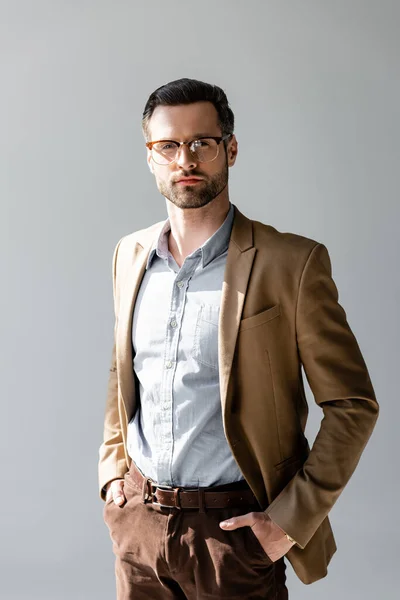 Homem de negócios bonito em óculos olhando para a câmera e de pé com as mãos em bolsos isolados em cinza — Fotografia de Stock
