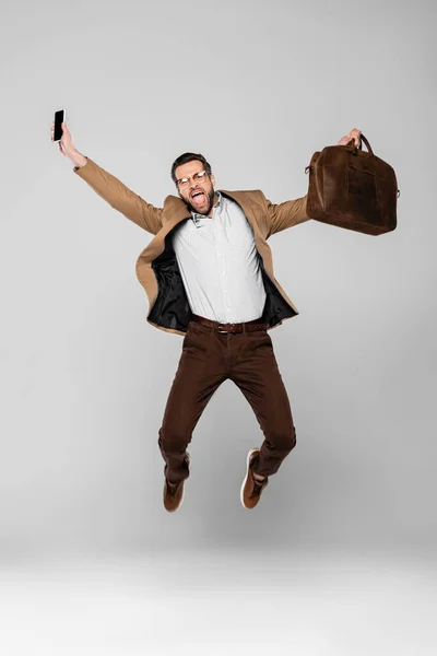 Aufgeregter Geschäftsmann hält Smartphone mit leerem Bildschirm und Tasche in der Hand, während er auf Grau schwebt — Stockfoto