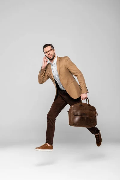 Hombre de negocios feliz hablando en el teléfono inteligente y bolsa de sujeción mientras levita en gris - foto de stock