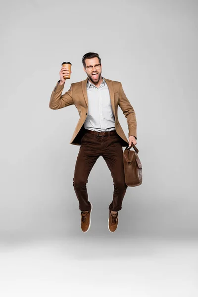 Feliz hombre de negocios sosteniendo bolsa y taza de papel mientras levita en gris - foto de stock