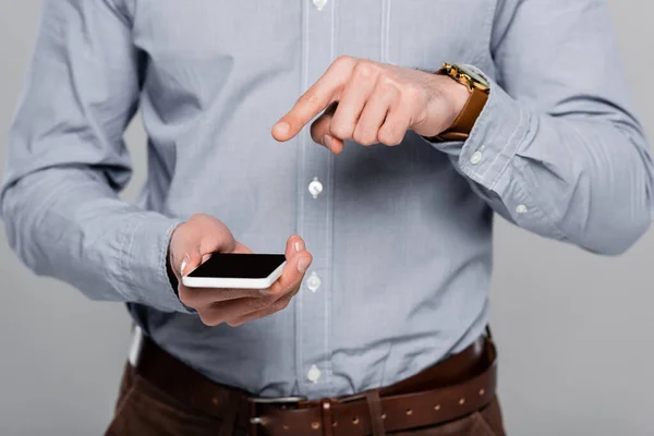 Vista recortada del hombre de negocios apuntando con el dedo al teléfono inteligente con pantalla en blanco aislado en gris - foto de stock