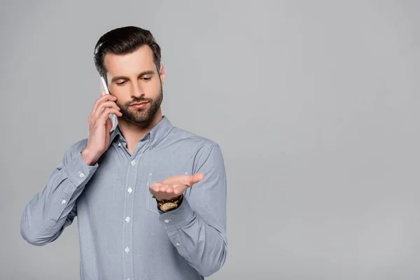 Homme d'affaires barbu pointant avec la main et parlant sur smartphone isolé sur gris — Photo de stock