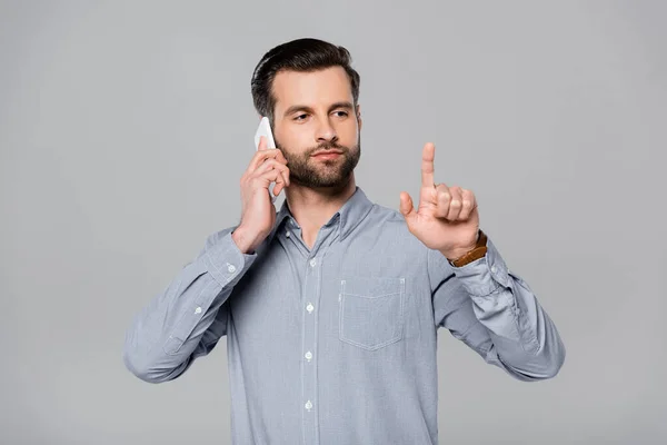 Hombre de negocios barbudo señalando con el dedo y hablando en el teléfono inteligente aislado en gris - foto de stock