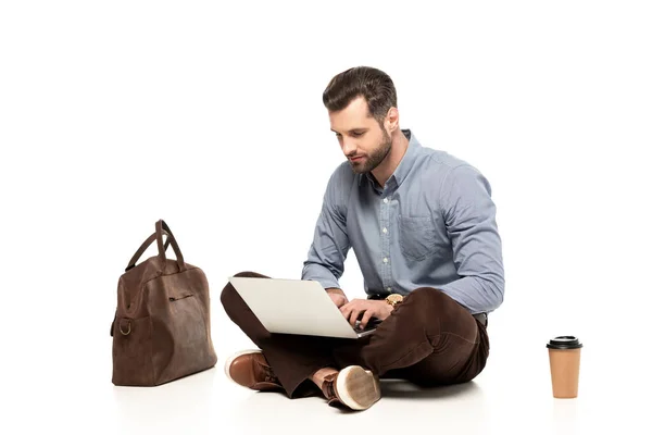 Handsome freelancer using laptop while sitting near bag and paper cup on white — Stock Photo