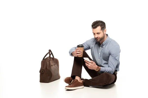 Bel homme utilisant smartphone tout en étant assis près du sac et tenant tasse en papier sur blanc — Photo de stock