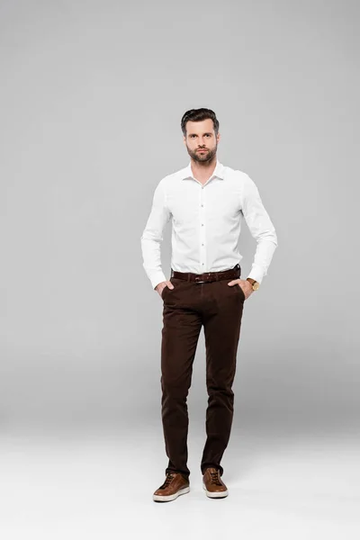 Бородатий бізнесмен в окулярах стоїть руками в кишенях на сірому — стокове фото