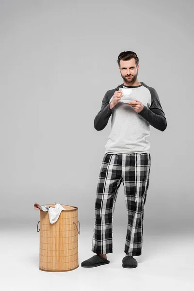 Homme barbu tenant une tasse de café près du panier à linge avec des vêtements sales sur gris — Photo de stock