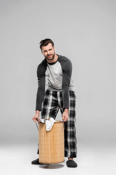 Homme barbu tenant panier à linge lourd avec des vêtements sales sur gris — Photo de stock