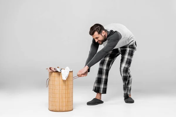 Emotional man pulling heavy laundry basket with dirty clothing on grey — Stock Photo