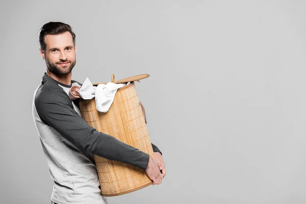 Homem bonito segurando cesta de roupa suja com roupas sujas isoladas em cinza — Fotografia de Stock