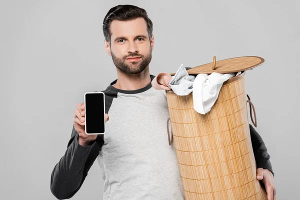 Homem bonito segurando cesta de lavanderia e smartphone com tela em branco isolado em cinza — Fotografia de Stock