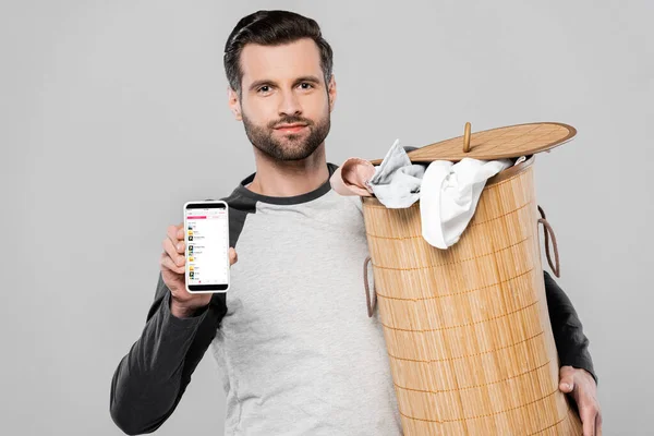 KIEW, UKRAINE - 23. MÄRZ 2020: schöner Mann mit Wäschekorb und Smartphone mit Apple-Musik-App auf dem Bildschirm isoliert auf grau — Stockfoto