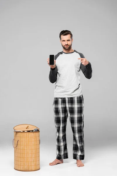 Bearded man pointing with finger at smartphone with blank screen near laundry basket on grey — Stock Photo
