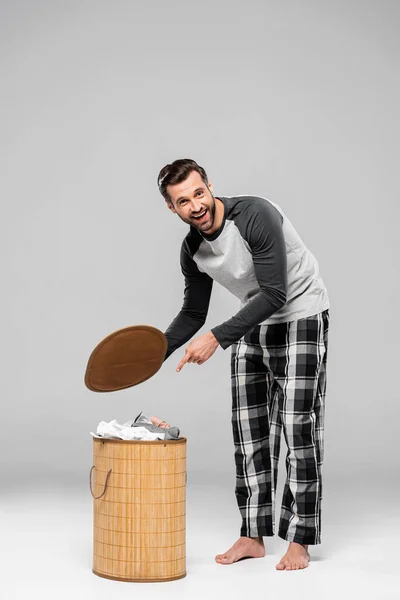 Uomo barbuto sorridente mentre punta con il dito al cesto della biancheria su grigio — Foto stock