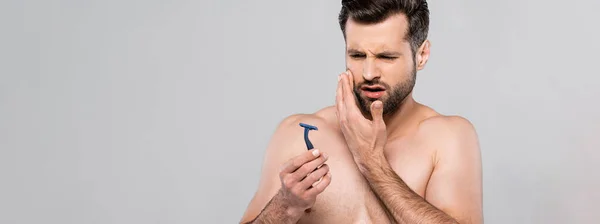 Panoramic shot of displeased man looking at razor isolated on grey — Stock Photo