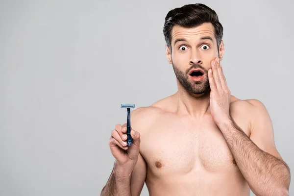 Hombre sorprendido y barbudo sosteniendo la navaja mientras tocaba la cara aislada en gris - foto de stock