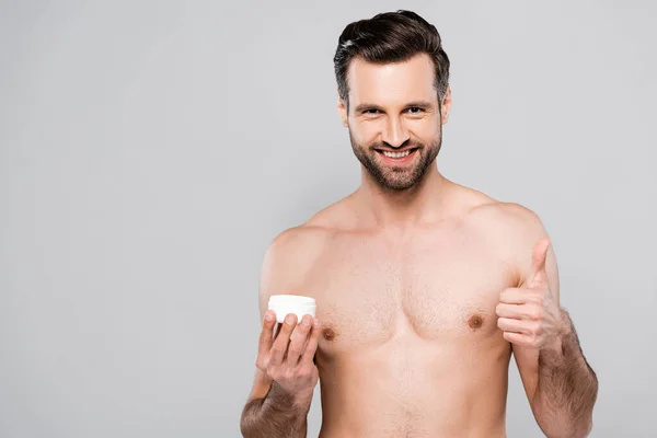Щасливий бородатий чоловік тримає тару з косметичним кремом і показує великий палець вгору ізольовано на сірому — стокове фото