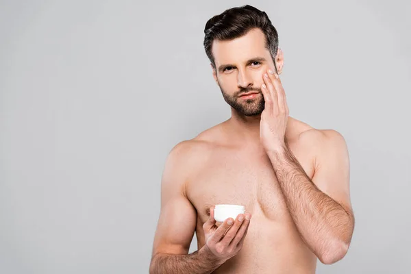 Hombre sin camisa que sostiene el envase mientras que aplica la crema cosmética aislada en gris - foto de stock