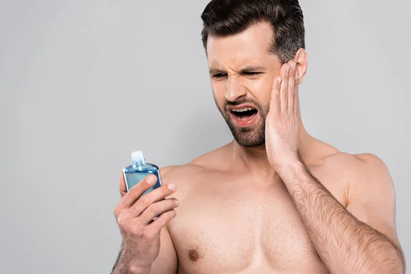 Hombre musculoso tocando la cara y mirando después de afeitarse loción aislada en gris - foto de stock