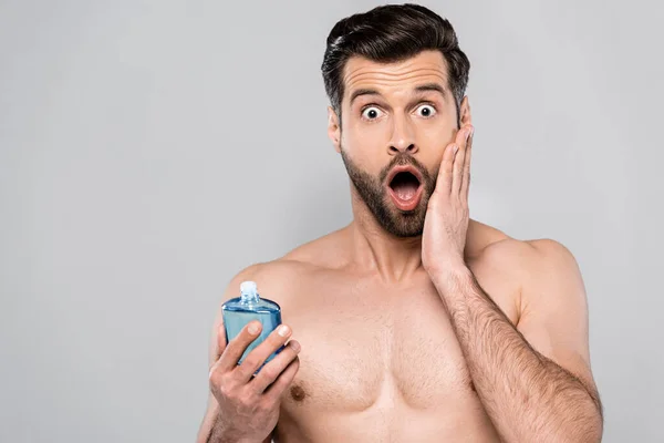Schockierter Mann hält Flasche mit After-Shave-Lotion und berührt Gesicht isoliert auf grau — Stockfoto