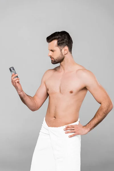 Hombre barbudo y musculoso en toalla mirando desodorante mientras está de pie con la mano en la cadera aislado en gris - foto de stock