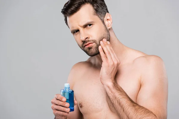 Handsome man holding bottle with after shave lotion and touching face isolated on grey — Stock Photo