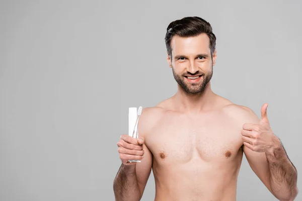 Homem feliz e muscular segurando pasta de dentes e escova de dentes enquanto mostra o polegar isolado no cinza — Fotografia de Stock