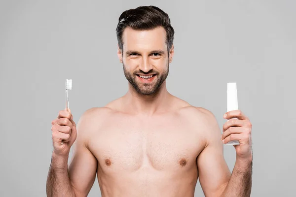 Homem feliz e muscular segurando pasta de dentes e escova de dentes isolado em cinza — Fotografia de Stock