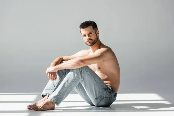 Sunlight on muscular man in blue jeans sitting on grey — Stock Photo