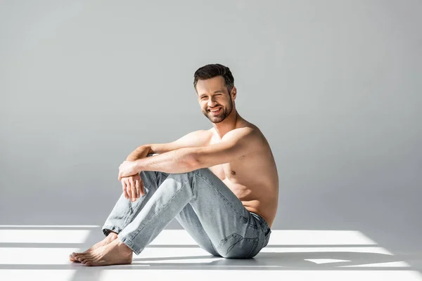 Sol en el hombre feliz y sin camisa en pantalones vaqueros azules sentados en gris - foto de stock