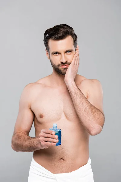 Muscular man holding bottle with blue after shave lotion and touching face isolated on grey — Stock Photo