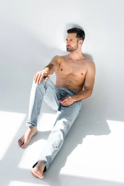 Sunshine on muscular man in blue denim jeans sitting on grey — Stock Photo