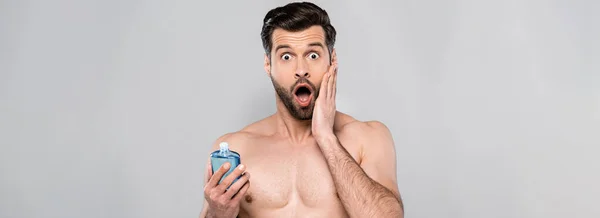 Панорамний знімок здивованого чоловіка торкається обличчя і тримає пляшку з синім після гоління лосьйону ізольовано на сірому — Stock Photo