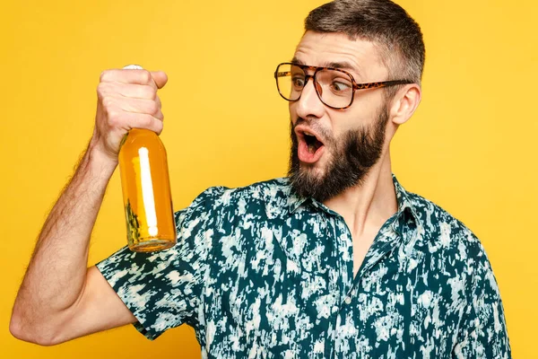 Шокований бородатий хлопець в окулярах дивиться на пляшку пива ізольовано на жовтому — стокове фото