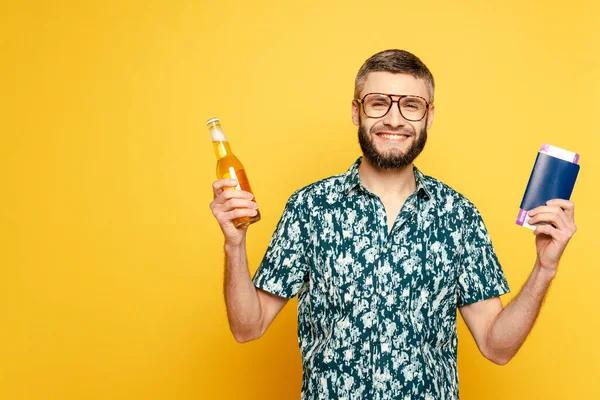 Smiling bearded guy in glasses with bottle of beer and air tickets in passport on yellow — Stock Photo