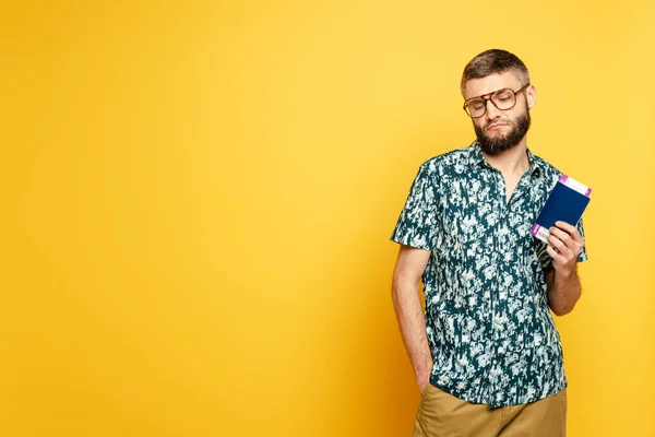 Sad bearded guy in glasses with air ticket and passport on yellow — Stock Photo