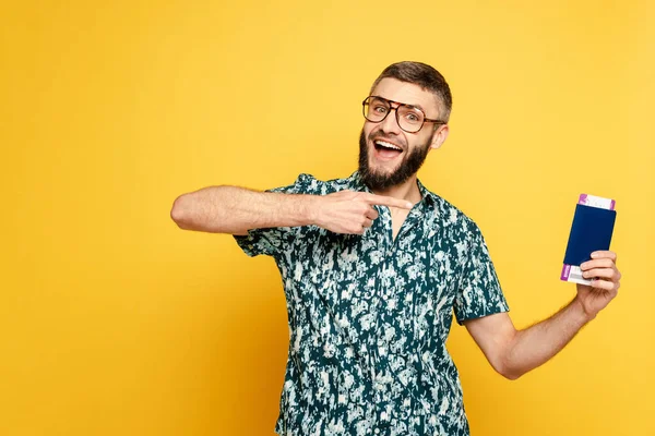 Glücklicher bärtiger Mann mit Brille zeigt auf Flugticket und Pass auf gelb — Stockfoto