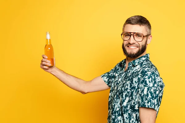 Vista laterale del ragazzo barbuto felice in bicchieri con bottiglia rinfrescante di birra sul giallo — Foto stock