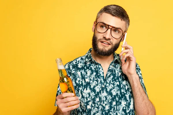 Pensive bearded guy in glasses with refreshing bottle of beer talking on smartphone isolated on yellow — Stock Photo