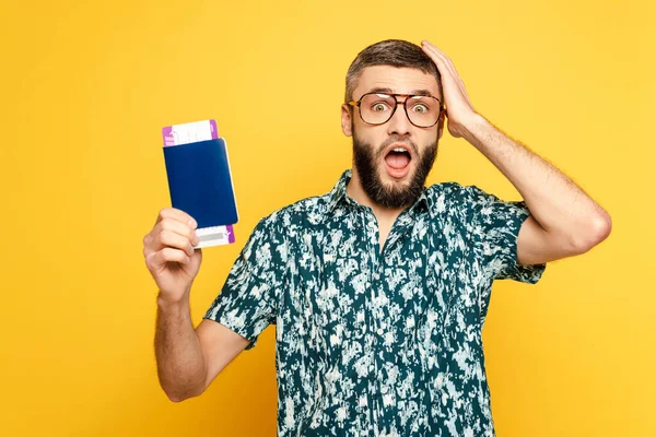Choqué barbu gars dans des lunettes avec passeport et billet tenant la tête sur jaune — Photo de stock