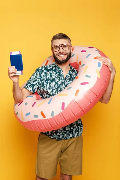 Smiling bearded guy in glasses with donut swim ring and passport on yellow — Stock Photo