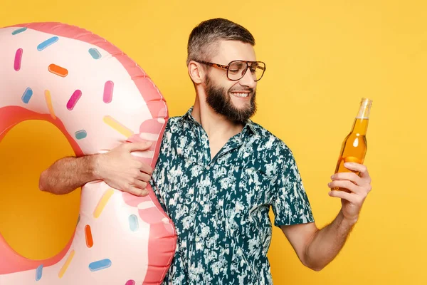 Ragazzo barbuto sorridente in occhiali con anello di nuoto ciambella e birra su giallo — Foto stock