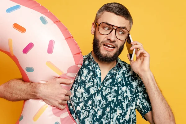 Confundido barbudo chico en gafas con anillo de natación donut hablando en el teléfono inteligente en amarillo - foto de stock