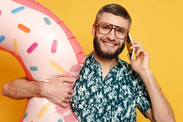 Sourire barbu gars dans des lunettes avec anneau de natation beignet parler sur smartphone sur jaune — Photo de stock